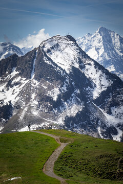 View of the Stoos mountain peak with a cross at the summit, snow-covered slopes, and a person standing on a grassy trail in Schwyz, Schwyz, Switzerland.