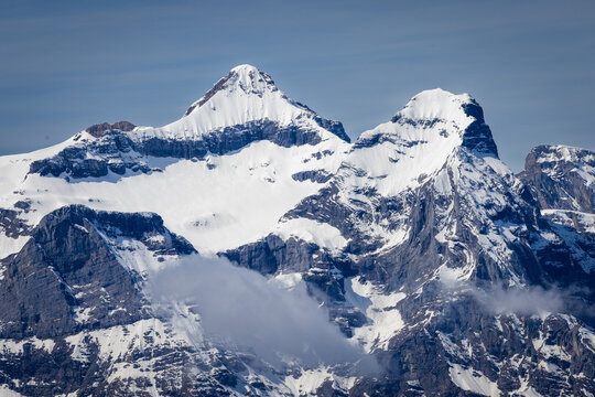 View of the snow-capped peaks of Stoos with rugged rocky cliffs and wispy clouds under a clear blue sky in Schwyz, Schwyz, Switzerland.