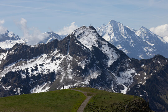 View of Stoos mountain landscape with snow-capped peaks, a hiking path in the green foreground, and a clear blue sky in Schwyz, Schwyz, Switzerland.
