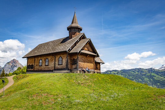 View of the Stoos chapel, a traditional wooden church situated on a lush green hill with people resting on a bench under a clear blue sky, Schwyz, Schwyz, Switzerland.