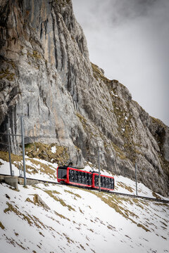 View of the red Mount Pilatus cogwheel railway train ascending a steep, snow-covered rocky mountain slope in Alpnach, Obwalden, Switzerland.