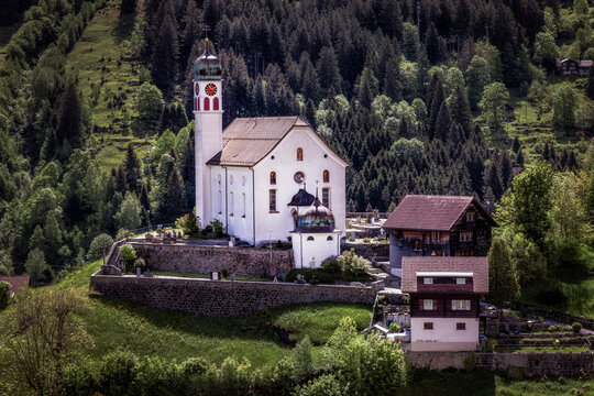 View of the iconic Wassen Church perched on a hill, surrounded by lush green slopes, dense pine forests, and traditional Swiss alpine architecture in Wassen, Uri, Switzerland.