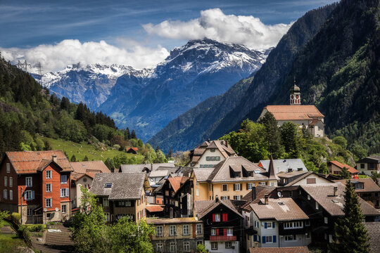 View of the historic village of Wassen with its prominent church perched on a hill, surrounded by lush green slopes and the snow-capped peaks of the Swiss Alps in Wassen, Uri, Switzerland.