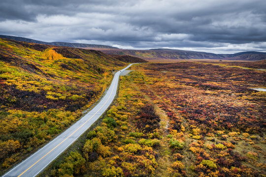 Aerial view of Richardson Highway winding through the vibrant autumn tundra of Alaska with colorful foliage and a dramatic cloudy sky in Salcha, Alaska, United States.