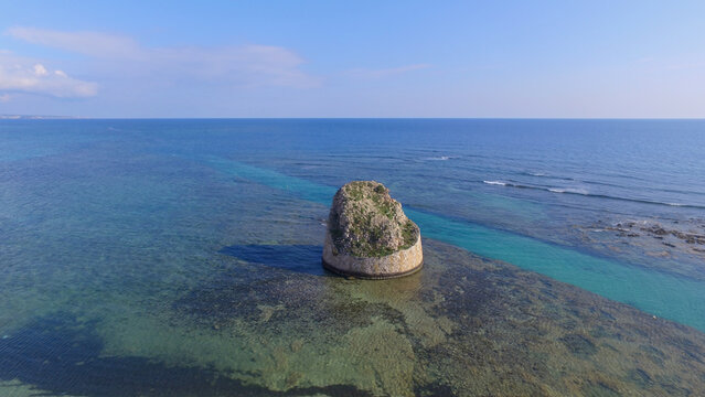 Aerial view of the historic Torre Pali watchtower standing in the clear turquoise waters of the Ionian Sea, surrounded by rocky shallows in Torre Pali, Apulia, Italy.