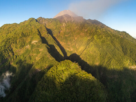 Aerial view of Mount Merapi with lush green slopes and volcanic smoke rising from the summit in Selo, Central Java, Indonesia.