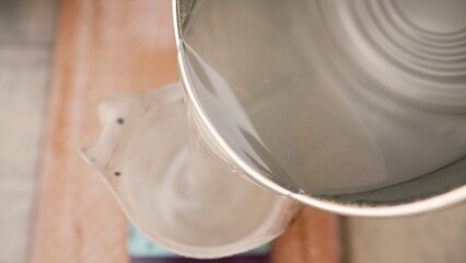 Obraz premium Overhead view of creamy white liquid flowing from metal bucket into ceramic vessel. Soft natural lighting, smooth texture, warm wooden background