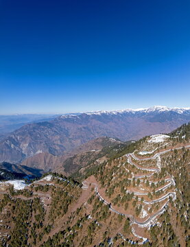 Aerial view of the winding mountain road leading to Pir Chinasi, surrounded by pine forests and snow-capped peaks in Muzaffarabad, Azad Jammu Kashmir, Pakistan.