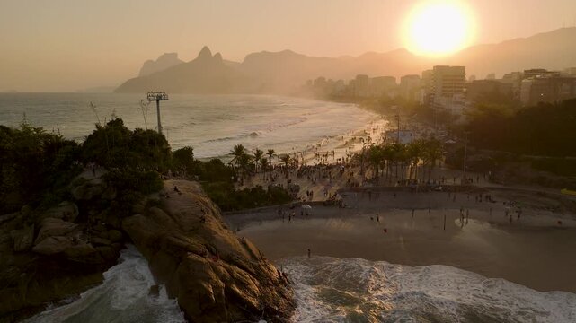 Aerial view of Arpoador rock formation at sunset with waves crashing against the shore and people gathered on the beach in Rio de Janeiro, State of Rio de Janeiro, Brazil.
