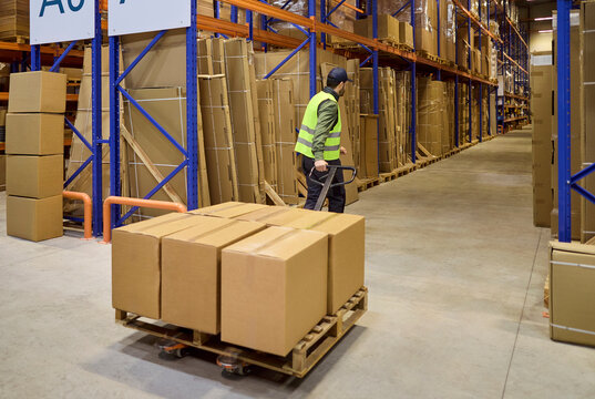 Warehouse worker moves pallet cartons on trolley. Organized storage logistics and shipping in a large center with high shelves and racking. Streamlined warehouse logistics and fast delivery.