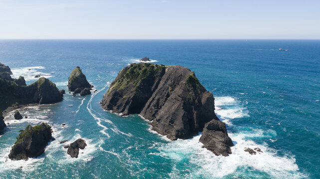 Aerial view of a rugged rocky island surrounded by turquoise and deep blue ocean waters with white sea foam crashing against the jagged cliffs in Japan.