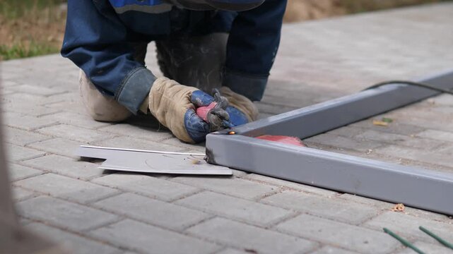 Kneeling worker joins gray metal beams on brick paving, with bright welding sparks and faint smoke rising at contact point during outdoor repair work in soft daylight, with welding, worker, kneeling