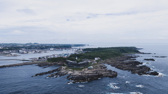 Aerial view of a rocky peninsula with a modern building, surrounded by the sea, with a coastal town and bridge in the background in Japan.