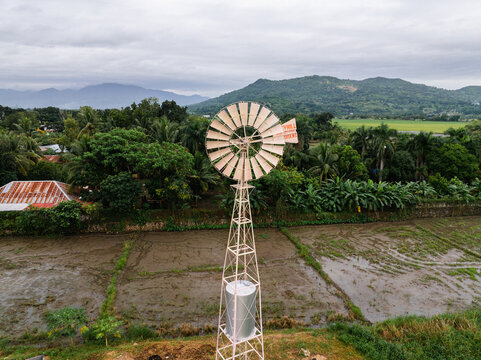 Aerial view of a metal windmill standing in a rural field surrounded by lush green trees and mountains under a cloudy sky in Bagabag, Cagayan Valley, Philippines.