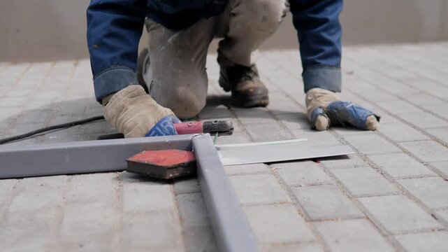 Construction worker kneels on light gray brick paving, fitting metal pieces with gloved hands and handheld tool, beside cable, dust, dark workwear, and muted outdoor industrial ground