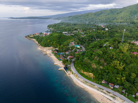 Aerial view of the coastal Santander - Barili - Toledo Rd winding through lush green hills and tropical vegetation along the shoreline in Alegria, Central Visayas, Philippines.
