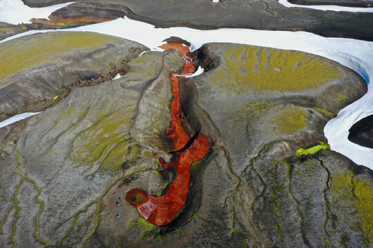Aerial view of Raudufossafjoll with its vibrant red river flowing through volcanic terrain, snow patches, and green moss, with people near the source in Hella, Rangarthing ytra, Iceland.