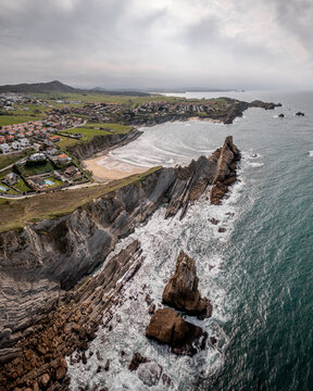 Aerial view of the dramatic rock formations and cliffs at Playa Del Portio, with the coastal town and sandy beach in the background, Liencres, Cantabria, Spain.