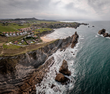 Aerial view of Playa Del Portio featuring dramatic jagged rock formations, crashing ocean waves, and the coastal town of Liencres with green fields in Liencres, Cantabria, Spain.
