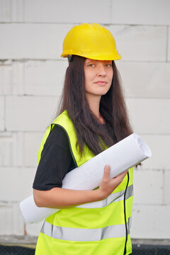 Young female construction worker in hi vis vest, helmet and gloves, carrying cable roll on her shoulder, bare new house wall near