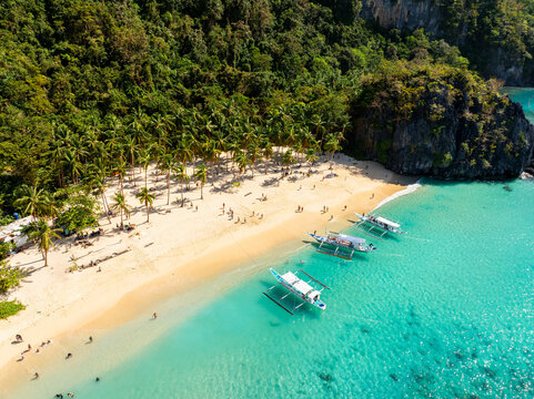 Aerial view of Seven Commandos Beach with turquoise waters, white sand, palm trees, limestone cliffs, and traditional outrigger boats in El Nido, Palawan, Philippines.