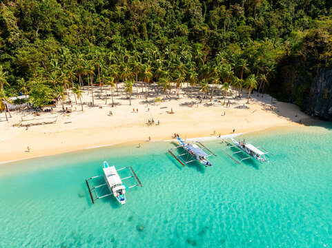 Aerial view of Seven Commandos Beach with white sand, lush palm trees, and traditional outrigger boats anchored in turquoise water in El Nido, Palawan, Philippines.
