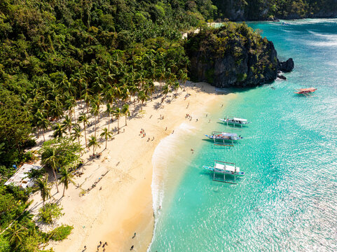 Aerial view of Seven Commandos Beach with turquoise water, sandy shore, palm trees, and traditional outrigger boats in El Nido, Palawan, Philippines.
