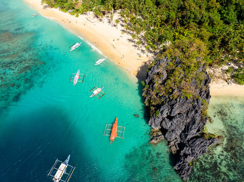 Aerial view of Seven Commandos Beach with turquoise water, traditional outrigger boats, and limestone rock formations in El Nido, Palawan, Philippines.