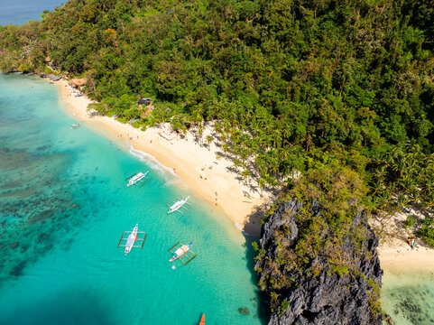 Aerial view of Seven Commandos Beach with turquoise water, traditional outrigger boats, white sand, and lush tropical forest in El Nido, Palawan, Philippines.