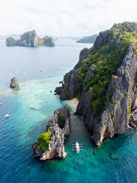 Aerial view of Shimizu Island with limestone cliffs, lush vegetation, and turquoise waters, featuring a traditional outrigger boat anchored in the bay in El Nido, MIMAROPA, Philippines.
