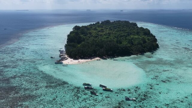 Aerial view of a lush tropical island surrounded by turquoise coral reefs and anchored boats in Karimunjawa, Central Java, Indonesia.