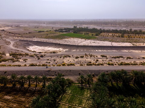 Aerial view of the arid landscape featuring lush date palm groves, green agricultural fields, and a long road stretching across the desert terrain in Panjgur, Balochistan, Pakistan.