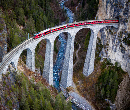 Aerial view of the Glacier Express train crossing the iconic stone Landwasser Viaduct above a river, surrounded by dense pine forests and rocky cliffs in Bergun Filisur, Graubunden, Switzerland.