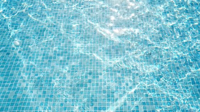 Top down slow motion view of pool water rippling over mosaic tile floor in sunlit shallow basin, shimmering reflections and patterns on blue surface