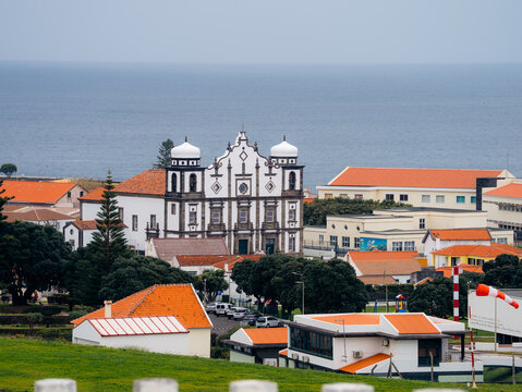 View of the Church of Nossa Senhora da Conceicao with orange-tiled roofs and lush trees overlooking the Atlantic Ocean in Santa Cruz das Flores, Azores, Portugal.