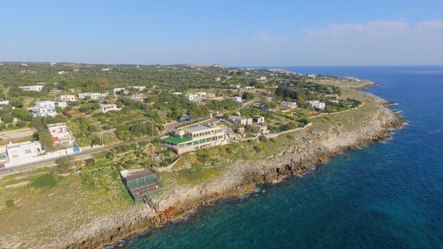 Aerial view of the rocky coastline and Mediterranean sea with scattered buildings and olive groves in Castrignano del Capo, Apulia, Italy.