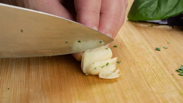 Woman slices fresh garlic cloves with a sharp knife on a wooden cutting board. Blade chops aromatic ingredients to prepare healthy food and add flavor to a culinary dish in a kitchen.
