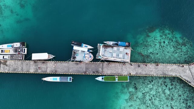 Aerial view of small boats moored at a concrete pier surrounded by clear turquoise water and coral reefs in Karimunjawa, Central Java, Indonesia.