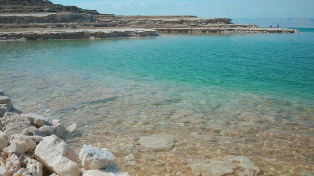 Calm clear turquoise water at shore of Dead Sea on Jordan side, salt covered layers of sand visible in distance, mountains far background are Israel