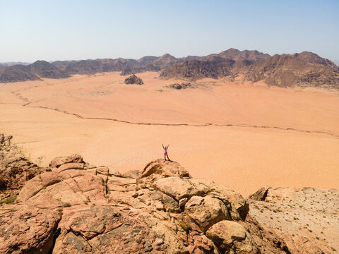 Aerial view of a person standing with arms raised on a rocky outcrop overlooking the vast desert landscape of the Wadi Rum Protected Area in Aqaba Governorate, Jordan.
