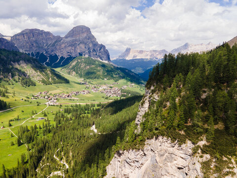 Aerial view of the village of Corvara nestled in a lush green valley surrounded by dramatic Dolomite peaks, pine forests, and rocky cliffs. Corvara, Trentino-Alto Adige, Italy.