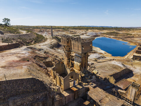 Aerial view of the abandoned Achada do Gamo mine site with industrial ruins, a tall chimney, and a blue water reservoir in an arid landscape in Achada do Gamo, Beja, Portugal.