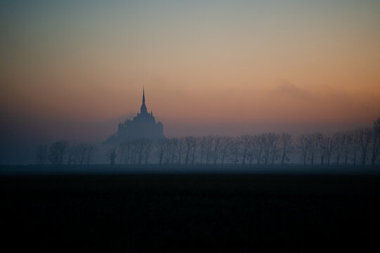 Aerial view of the silhouette of Mont Saint-Michel abbey rising above the mist at dawn with a row of trees in the foreground in Mont Saint-Michel, Normandy, France.
