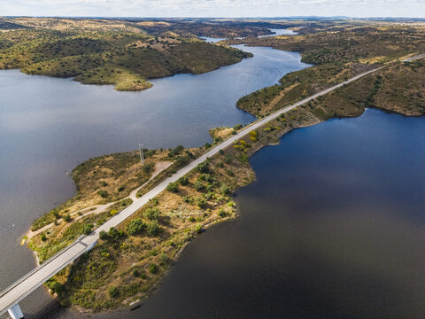 Aerial view of Albufeira do Alqueva reservoir with a long bridge crossing the water, surrounded by rolling hills and dry vegetation under a clear sky Alqueva, Distrito de Evora, Portugal.