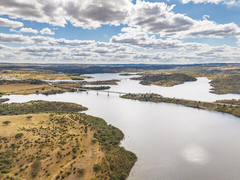 Aerial view of Albufeira do Alqueva reservoir with a long bridge crossing the water, surrounded by rolling hills, olive groves, and a cloudy sky in Alqueva, Distrito de Evora, Portugal.