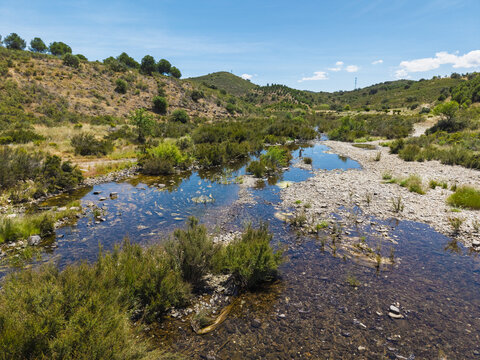 Aerial view of a shallow stream flowing through the rural landscape with rolling hills, green vegetation, and rocky banks in Vaqueiros, Faro District, Portugal.