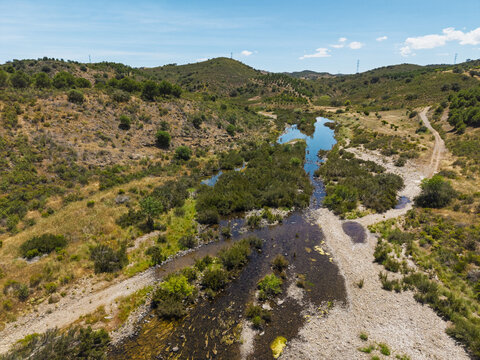 Aerial view of a winding river flowing through the dry, hilly landscape of Vaqueiros, with sparse vegetation and dirt paths in Vaqueiros, Faro District, Portugal.