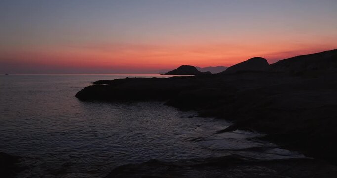 Soft orange and red light up the pre dawn morning as the sun rises over the island of Milos, Greece. Aegean sea water gently flows to the shore of Mandrakia looking toward Sarakiniko beach. Quiet 