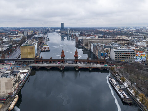 Aerial view of the historic red-brick Oberbaum Bridge spanning the Spree River, surrounded by urban architecture and river traffic under a cloudy winter sky in Berlin, Berlin, Germany.