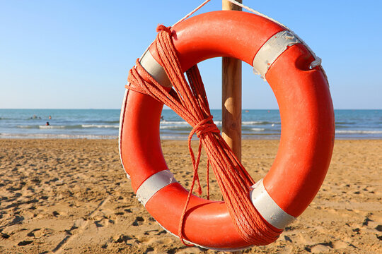 Orange lifebuoy with rope hanging on wooden pole on sandy beach near sea under clear sky